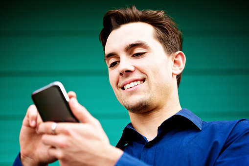 A handsome young man smiles, looking down in delight at an image on his smartphone. Low-angle view.