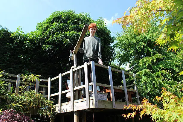 Photo showing a young teenage boy standing on his treehouse decking platform, overlooking the garden below.  The platform is edged with a simple picket-style fence, for safety reasons.  He is pictured stands next to a 'crane' device, used to raise baskets of food up to the treehouse.