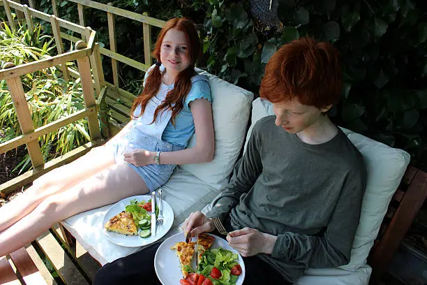 Photo showing children (boy and girl / brother and sister) enjoying an alfresco breakfast, eating outside on the deck / decking on their elevated treehouse.  They are enjoying the shade on a sunny morning, whilst looking at the garden below.