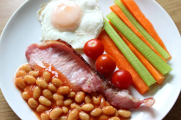 Photo showing a healthy full English fried breakfast, consisting of a rasher of grilled bacon, baked beans (reduced sugar and salt), cherry tomatoes, a dry fried / poached egg and julienne slices of carrots and celery.  This is a low calorie version of a traditional fried breakfast meal, served up as part of a healthy eating diet plan.