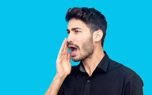 Profile view portrait of young man shouting against blue background. Young man putting his hand near his mouth and screaming loudly with serious facial expression. His hand covering his mouth. Horizontal composition. Image taken in studio isolated on blue and edited from Raw format. Shouting young man wearing a black button down shirt and standing in front of blue background. He is in 20s. Young man has got short,black hair and beard. His ethnicity belongs to Turkish  -  middle eastern ethincity. Focus on young man's face.