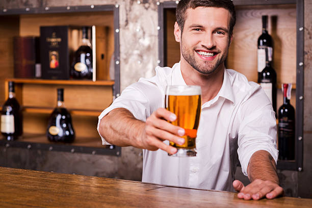 Let me quench your thirst! Happy young male bartender in white shirt stretching out glass with beer and smiling while standing at the bar counter quench your thirst pictures stock pictures, royalty-free photos & images