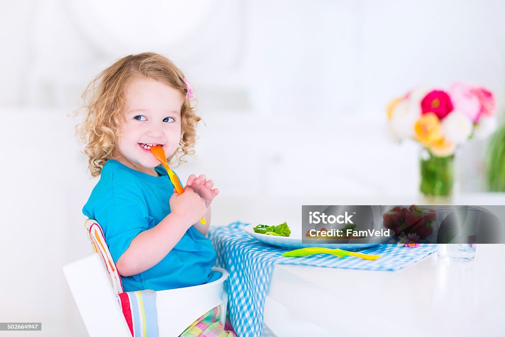 Little happy girl eating salad for lunch Happy little girl, cute curly toddler, eating fresh vegetables for lunch, healthy salad snack, corn, broccoli, carrots and strawberry fruit in a white dining room sitting in a high chair Healthy Lifestyle Stock Photo
