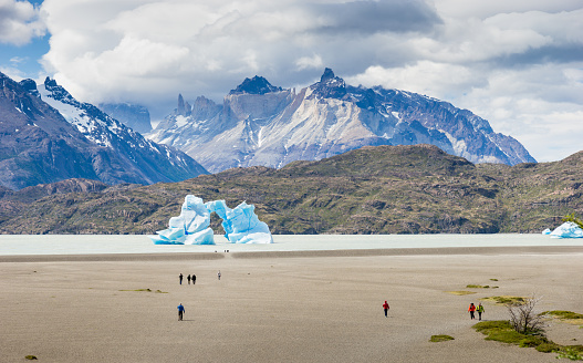 Torres del Paine, Chile - November 4, 2015: Tourists are walking on the shore of Lago Grey, admiring Cuernos del Paine clearly visible in the background. Arch shaped ice float is visible in the center of the photograph.