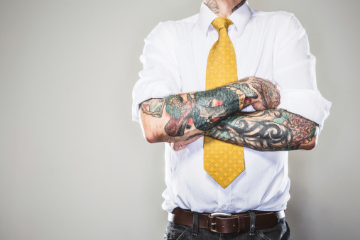 A business man stands with his tattooed arms folded across his white collared shirt and tie.  Two forearm sleeve tattoos.  Representing a new generation of modern business standards and style.