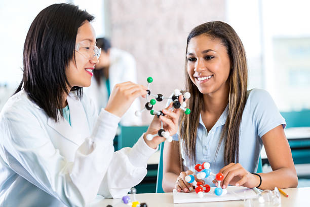 Young Asian female teacher helps teenage private high school student study molecular structure. The teacher is holding a plastic educational model. The teenage girl is interested as her teacher talks. The student is wearing a school uniform and the teacher is wearing a white lab coat.