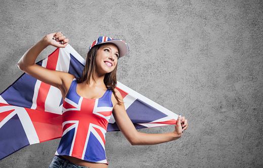 Cheerful cute English girl holding a flag, wearing a tank top and a cap with Union Jack flag, patriotism and enjoyment concept