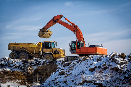 Winter scene with excavator loading earth to large yellow dump truck in road construction