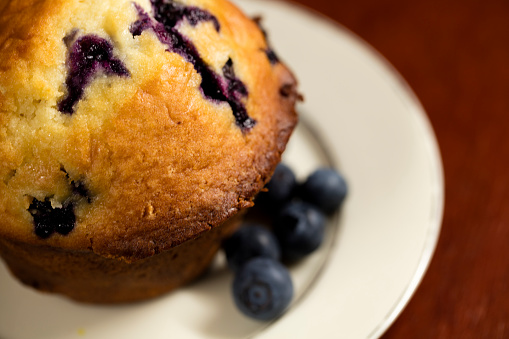 Freshly baked blueberry muffin on a white plate with fresh blueberries on the side of the muffin