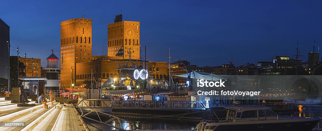 Oslo Aker Brgge waterfront bars restaurants illuminated at dusk Norway The iconic Functionalist towers of Oslo City Hall, Radhus, spotlit against the chrome blue dusk sky overlooking the crowded promenades and marinas of Aker Brygge, the popular leisure, dining and exclusive residential district in the heart of downtown Oslo, Norway's picturesque capital city. ProPhoto RGB profile for maximum color fidelity and gamut. Oslo Stock Photo