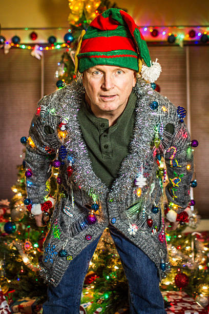 Ugly Sweater Elf A Caucasian man wearing an ugly cardigan holiday sweater, and an elf hat, with a decorated tree in the background. christmas ugliness sweater nerd stock pictures, royalty-free photos & images