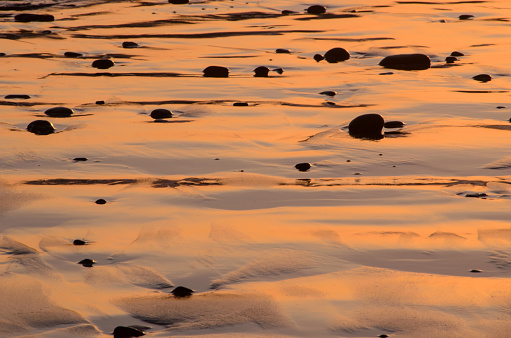 An abstract full frame close up photo of pebble silhouettes on wet beach sand that is reflecting the bright orange and pink colors of sunset