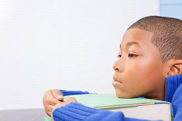 Exclusion. Sad, African descent school boy with chin on book. Depression, exclusion, left behind.  Sad African descent boy with chin on textbook at school.  school exclusion stock pictures, royalty-free photos & images