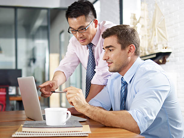 asian and caucasian businessmen working together in office asian and caucasian business executives looking at laptop screen while having a discussion in a multinational company. expatriate photos stock pictures, royalty-free photos & images