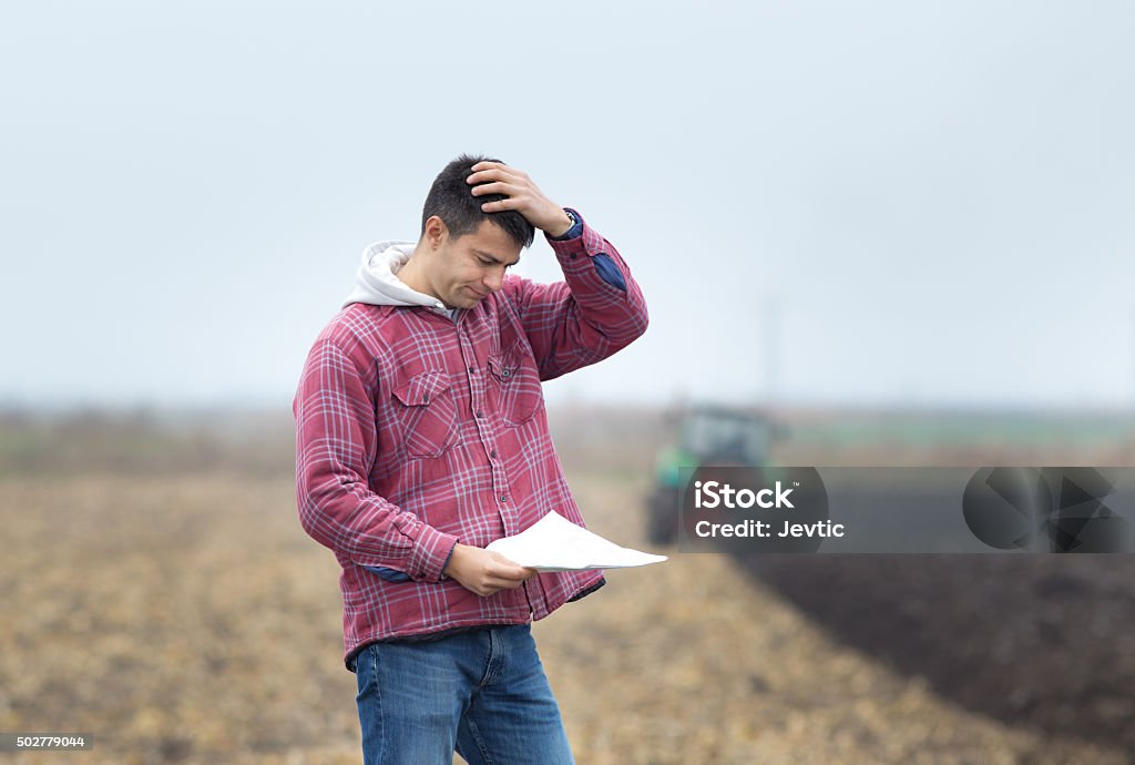 Worried man on the field Worried young farmer standing on field and  looking at papers from bank, tractor in background Farmer Stock Photo