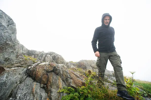 A mature man wearing hiking clothes and boots walks on a rocky mountain summit the mist. Shot at Grimselpass in the Swiss Alps.