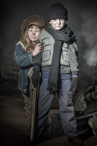 Two homeless kids in old clothes in slum. Serious little boy in a black cap is holding a wooden stick. Cheerful red-haired little girl in big cap peeking out from behind the boy. Staged in an abandoned factory