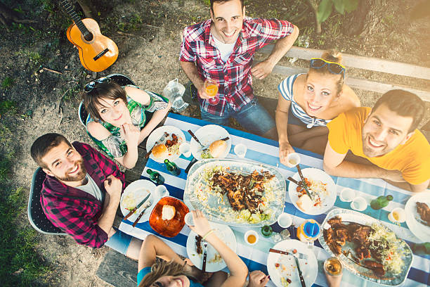 Group of people having lunch outdoors. Group of young adults having lunch outdoors. Sitting at wooden table and eating barbecue, drinking beer,wine and soft drinks. Sun is low and entering the frame from the right hand side. Each person is looking at camera and smiling. Top view. restaurant place setting dinner dinner party stock pictures, royalty-free photos & images