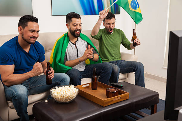 Guys cheering for Brazil on TV Young Latin men cheering for Brazil on TV while hanging out and drinking some beer at home excited football fan cheering for his team on tv stock pictures, royalty-free photos & images