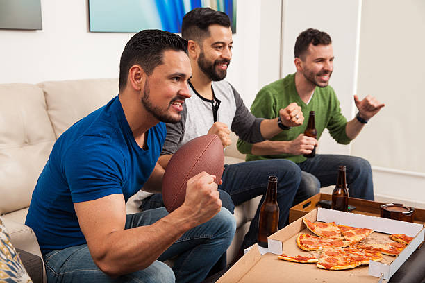 Friends watching american football Profile view of a group of male friends cheering for their football team while watching the game at home excited football fan cheering for his team on tv stock pictures, royalty-free photos & images