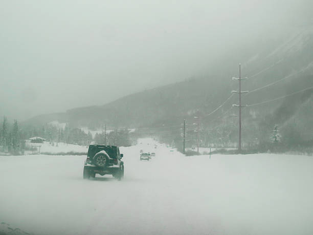 A Jeep and a line of vehicles navigate a snow covered road in Colorado, north of Durango.  The road is completely covered in snow.  The line of cars is driving slowly on the slippery road conditions.