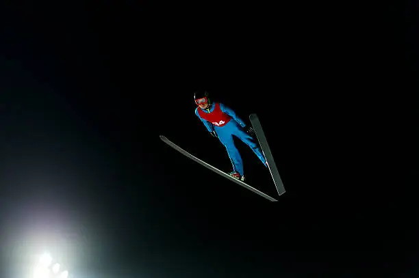 Front view of young male ski jumper during the long ski jump against the dark background, night shot against the light