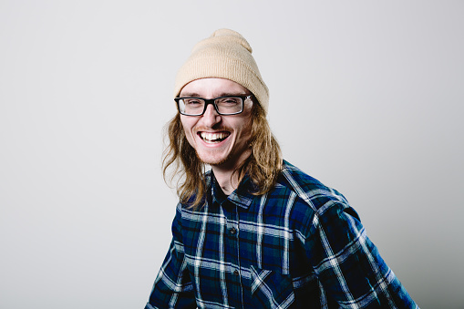 Studio portrait of a real man in his late 20s with long hair and glasses laughing and smiling looking at the camera. This image offers much copy space with room for your branding, logo or text.
