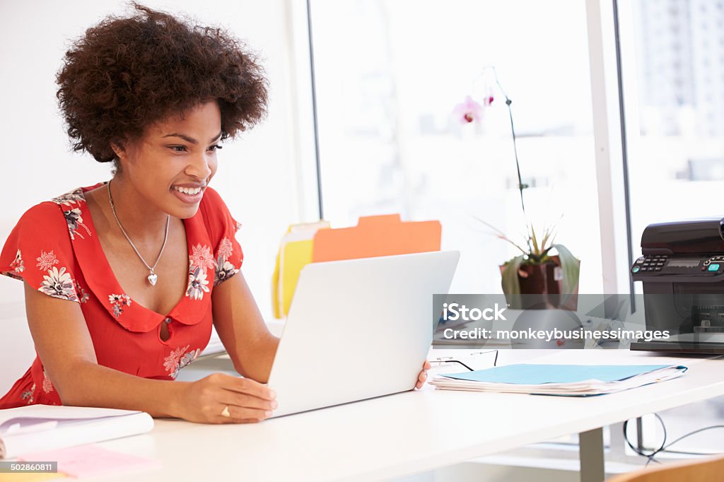 Smiling woman looking at silver laptop in studio Woman in office with laptop computer, files and paper on a white desk.  The African American woman has curly, dark-brown hair, a red v-neck shirt with a floral print, and a silver heart-shaped necklace draped around her neck.  The woman is smiling and looking at a silver laptop computer that is placed on a white desk.  A blue file folder, glasses, white paper and a pink memo sheet are also on the desk.  In the background, a black printer, green plant, white papers and files are on a table next to a line of office windows. 20-29 Years Stock Photo