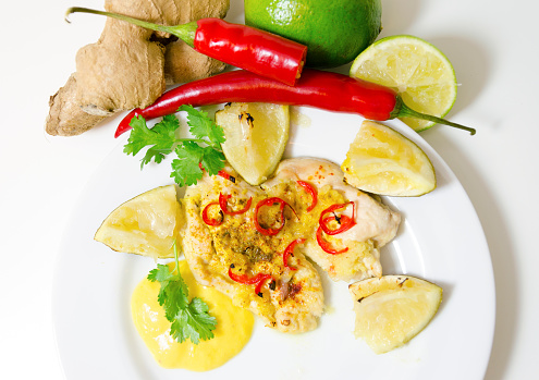 Top view of a roasted in the oven chicken breast fillet garnished with chili, ginger and lime served in a plate with mango sauce, raw vegetables on a white background. A healthy asian cuisine dish.
