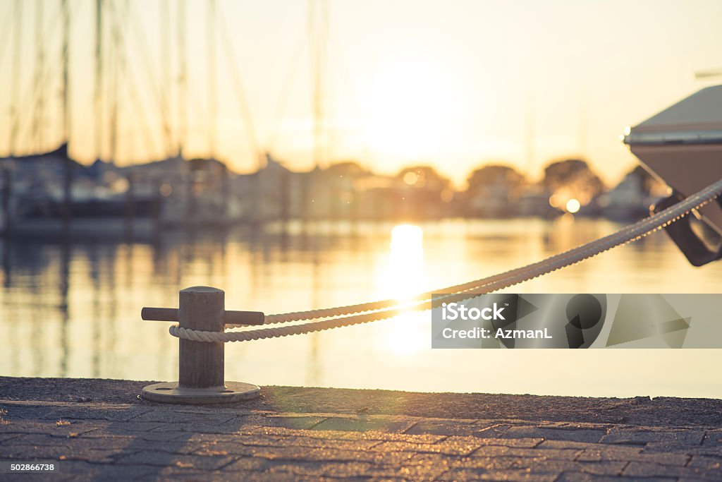 Yacht moored on harbour Yacht in port tied with a rope on mooring post on harbour during sunset. Sun and reflection of it in back. Nautical Vessel Stock Photo