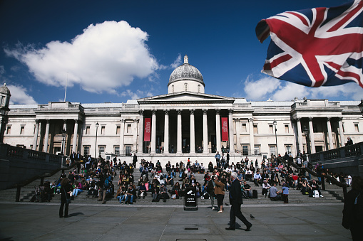 London, United Kingdom - April 14, 2014: Tourists are sitting on steps on Trafalgar Square, London. Trafalgar Square is a public square in the City of  Westminster, Central London, built around the area formerly known as Charing Cross. Surrounding the square  are the National Gallery ton the the north side and St Martin-in-the-Fields Church to the east.
