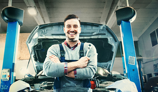 Wide angle portrait of smiling mid 30's car mechanic standing in front of a vehicle he's attending to. Holding a wrench in his hand. Car jack hydraulics in background. Low angle view.