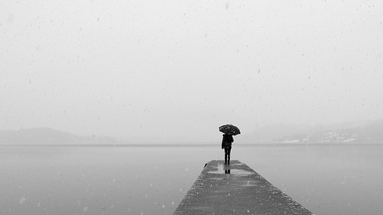 Lonely woman with umbrella standing on the edge of a pier a cold winter day with snow and rain.