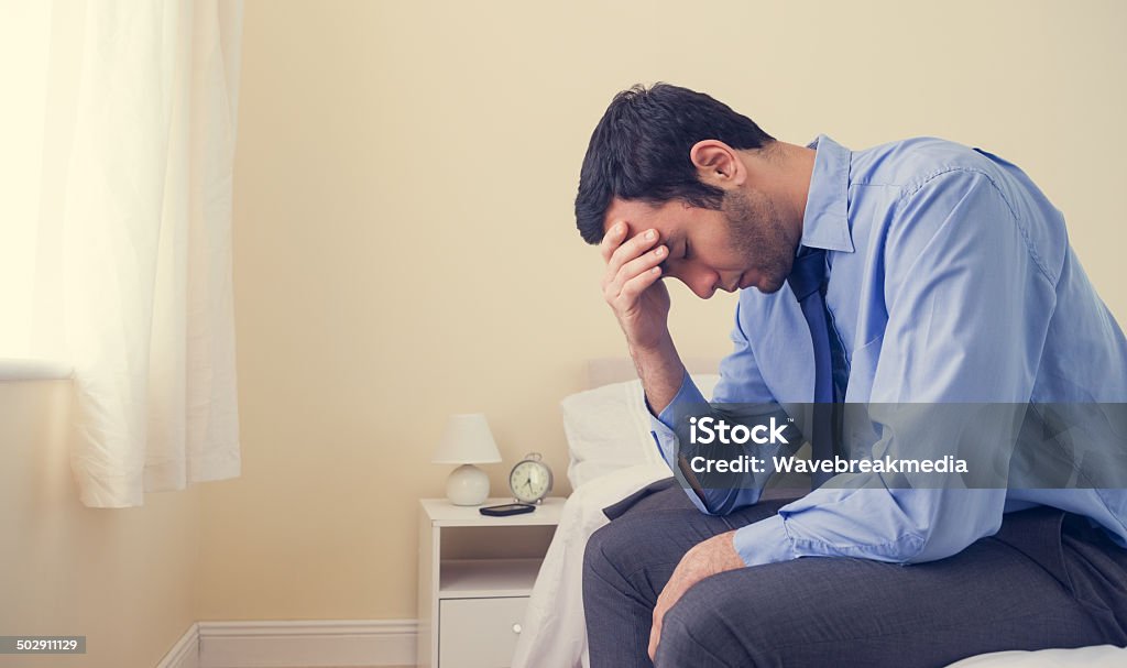 Depressed man sitting head in hands on his bed Depressed man sitting head in hands on his bed in a bedroom at home Blue Stock Photo