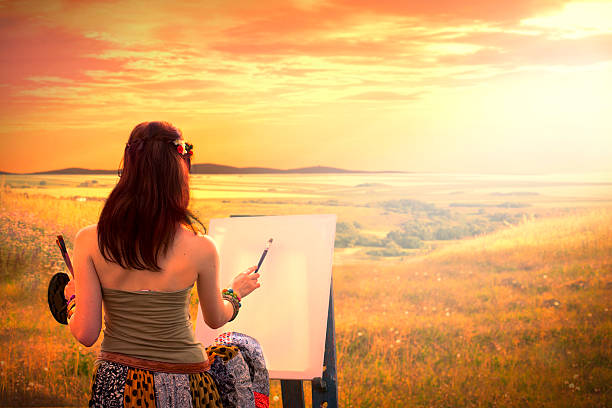 Senior Woman Sitting At Outdoor Table Painting Landscape By Lake. Smiling To Camera