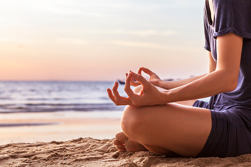 Young woman relaxing by practicing yoga on the beach at sunset, close-up of hands, gyan mudra and lotus position