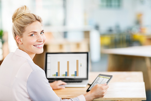 Rear view portrait of female entrepreneur using digital tablet and laptop at desk in office. Horizontal shot.