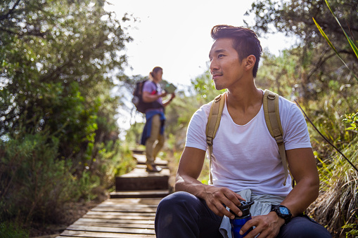 A photo of thoughtful male hiker looking away. He is sitting by boardwalk while friend is standing in background. They are in casuals. Active men are hiking in forest during weekends.