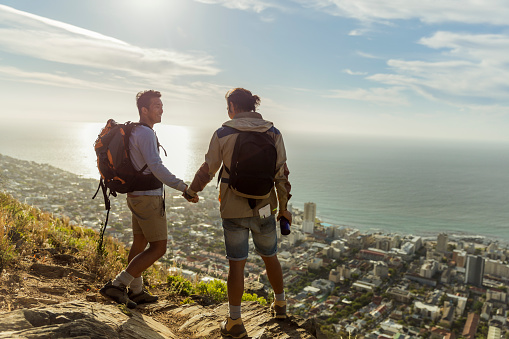 A photo of gay hikers holding hands while standing on rocks. Happy couple are carrying backpacks. Full length of young males are in casuals against sky.