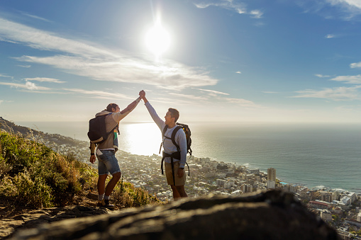 A photo of gay hikers celebrating success on hill. Happy couple are holding hands. Full length of young males are in casuals against sky.