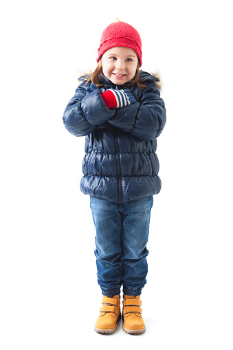 Studio full length portrait of cute 4 years old girl standing and posing with crossed arms isolated on white background. She is wearing winter clothes, red hat, colorful gloves, blue winter jacket and jeans.