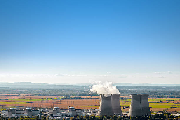 Horizontal composition photography with large copy space on clear blue sky background of french nuclear power station with four steaming cooling towers in countryside plain with smoke cloud (water condensation). Image taken from high angle view in Bugey, in Ain on the border of Isere department, Rhone-Alpes region in France (Europe). The nuclear power station is located at the extreme bottom of the image, in the middle of a plain landscape in France, near Lyon city. Shot in sunny day autumn season with green and brown field in countryside.