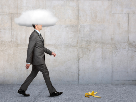 A banana peel in the path of a businessman walking on a sidewalk with his head in a cloud.