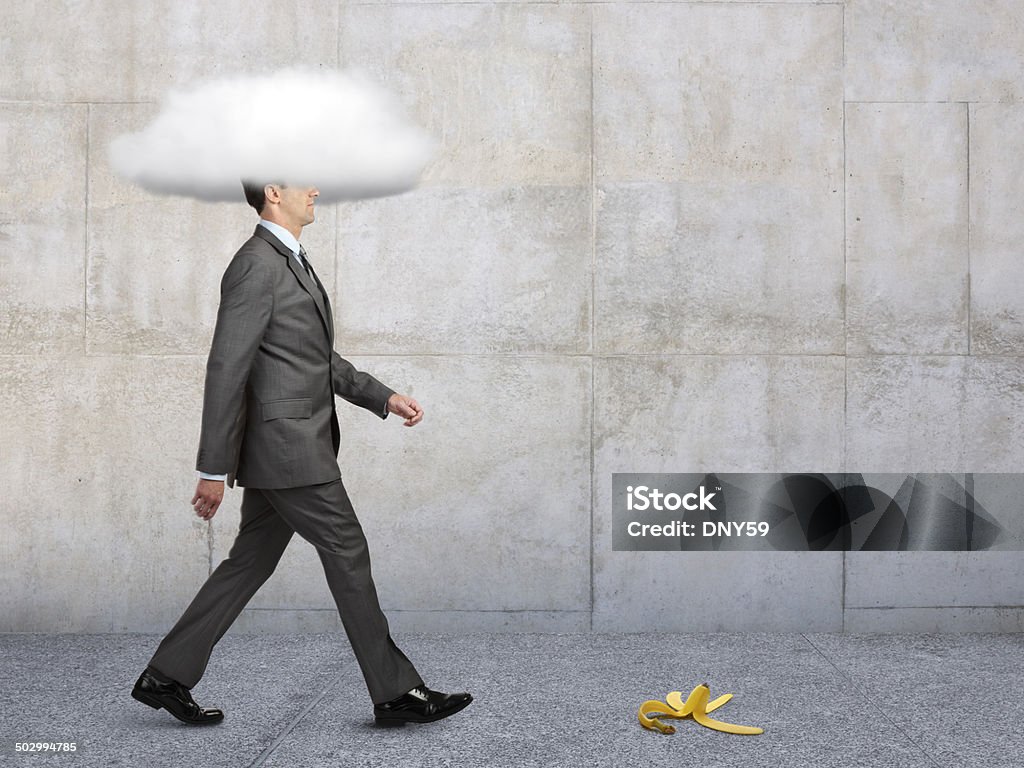 Businessman With Head In Clouds With Banana Peel In Path A banana peel in the path of a businessman walking on a sidewalk with his head in a cloud. Ignorance Stock Photo