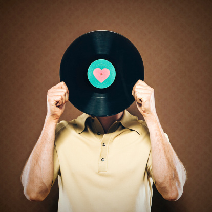 A man holds a 33⅓ rpm microgroove vinyl LP record up to his face as if it were his head, a heart shape in the middle of the record's label.  Retro wallpaper background, square crop.