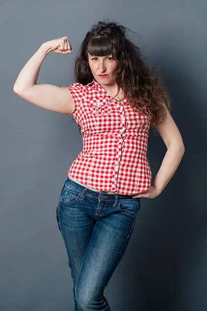 muscle concept - proud young woman with tomboy retro look raising her arm to enjoy female power, symbol of freedom,studio shot