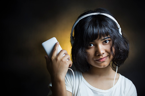 Indoor studio shot of a cute teenage girl listening music through headphones plugged in smart phone while standing against dark studio background with a golden glow behind her head and smart phone she is showing and looking at camera with smile. One person, head and shoulders, horizontal composition with copy space and selective focus.