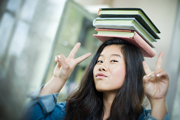 Happy Asian girl student balancing stack of books on head.