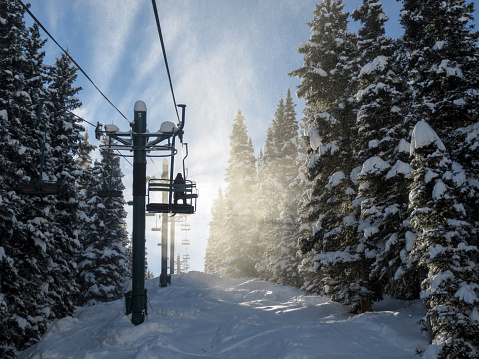 Image of the silhouette of a skier sitting on a chair lift, back lit by blowing snow.  The chair is ascending through snow covered pine trees.  The snow in the wind on this bright day appears white against blue sky.