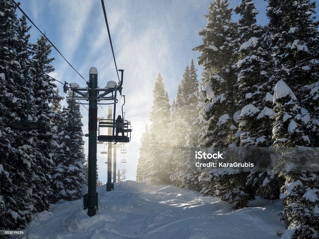 Silhouette of a skier and chair lift in blowing snow Image of the silhouette of a skier sitting on a chair lift, back lit by blowing snow.  The chair is ascending through snow covered pine trees.  The snow in the wind on this bright day appears white against blue sky. Durango - Colorado Stock Photo